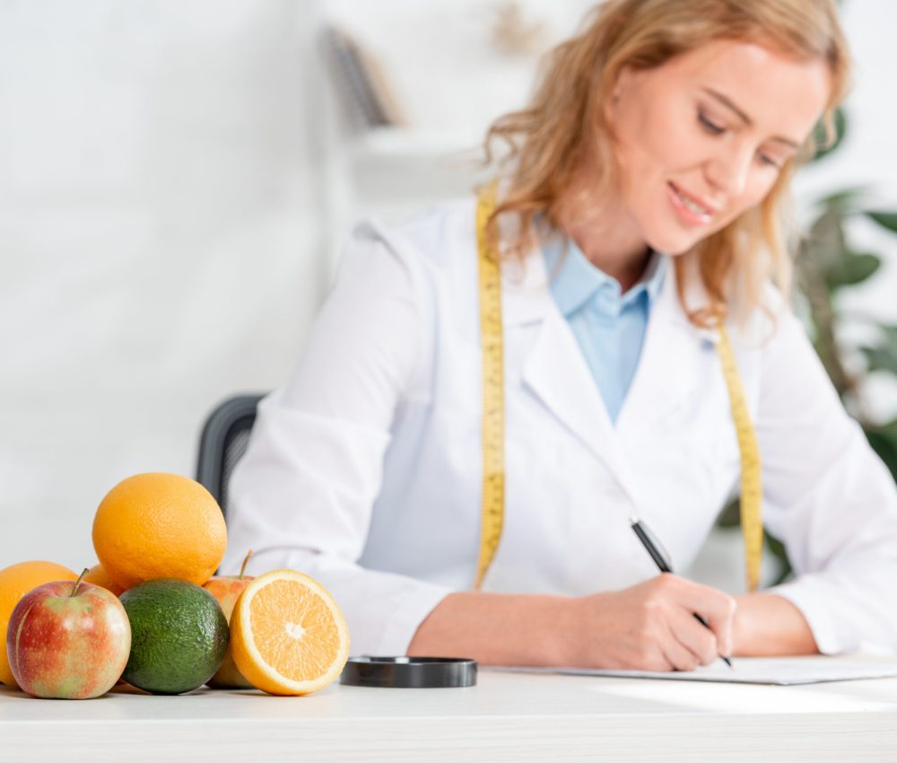 selective focus of fruits and vegetable on table and nutritionist sitting at table and writing on